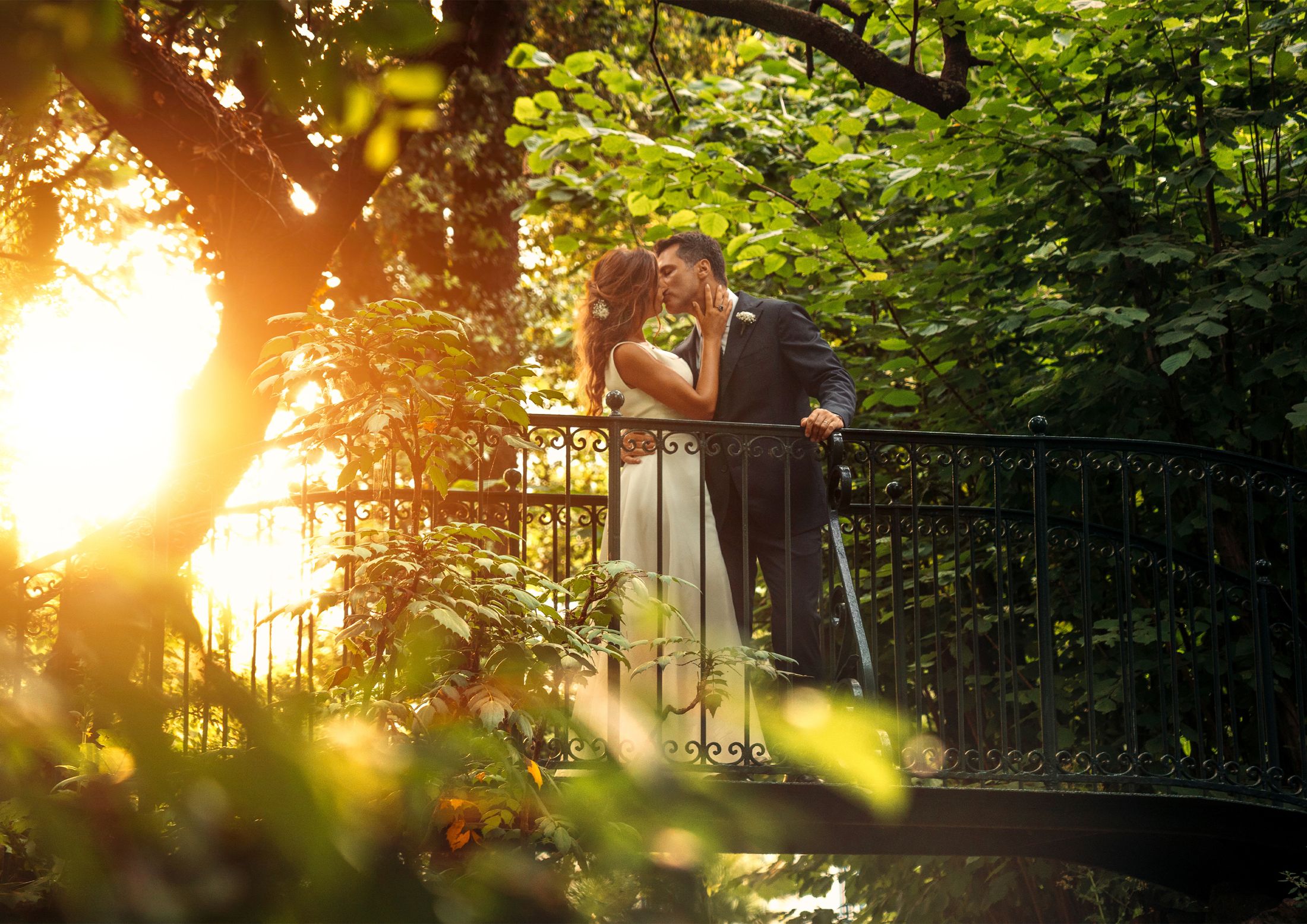 Bacio degli sposi su un ponticello in ferro battuto, controluce dorato tra gli alberi